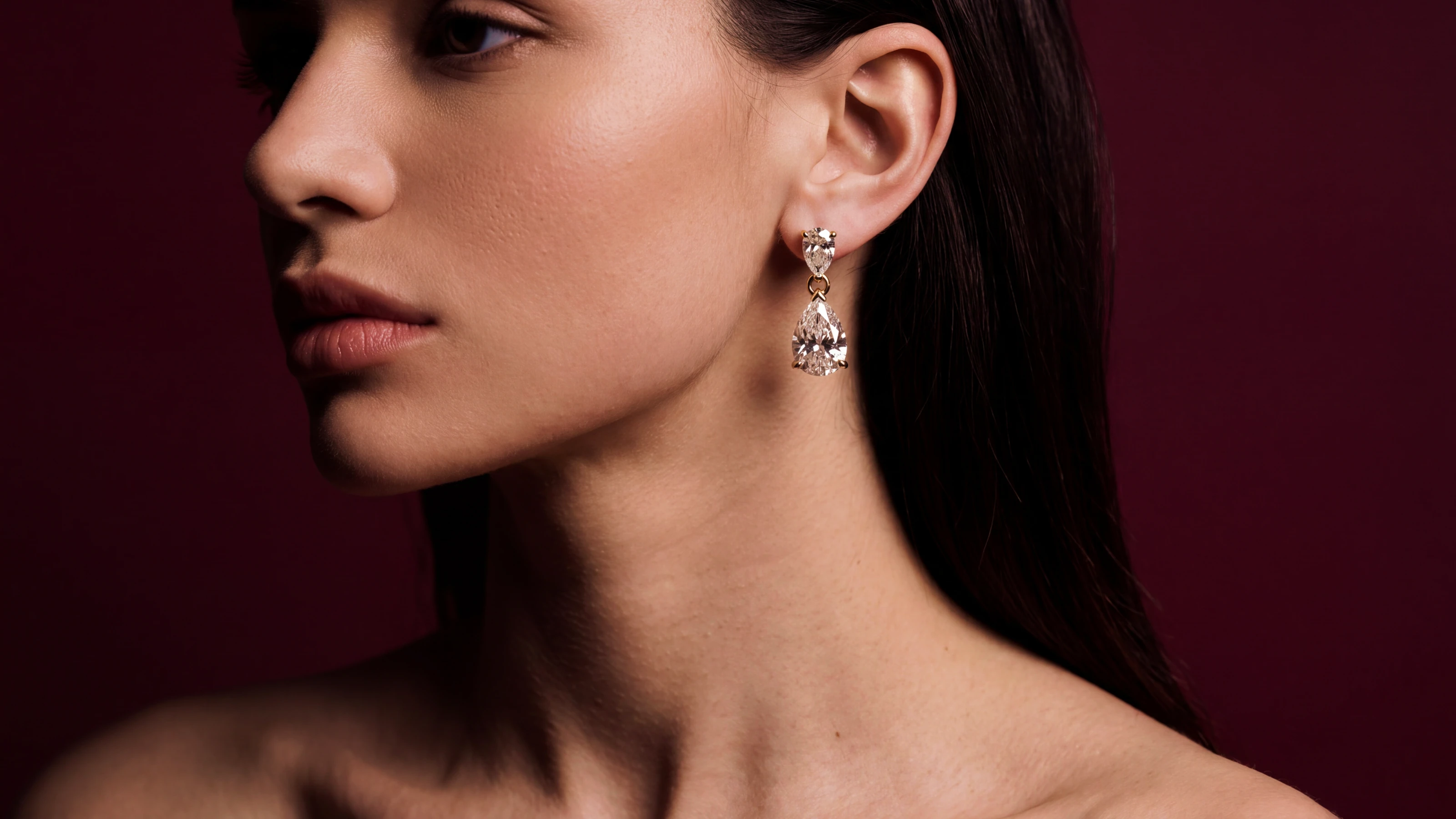 A model wearing diamond drop earrings against a burgundy background.